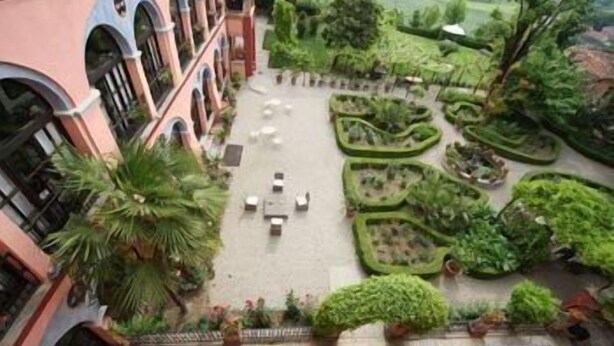 Un patio con una zona de estar central, rodeado de follaje y un edificio con ventanas de arco.