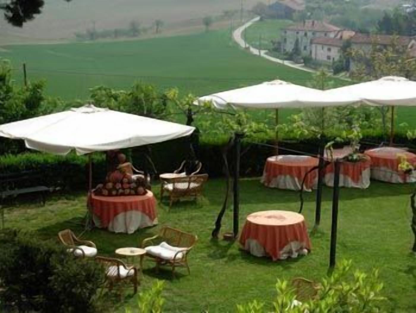 Ein Sitzbereich im Freien mit runden Tischen und Stühlen, beschattet von weißen Sonnenschirmen, mit Blick auf ein grünes Feld und ein Gebäude.