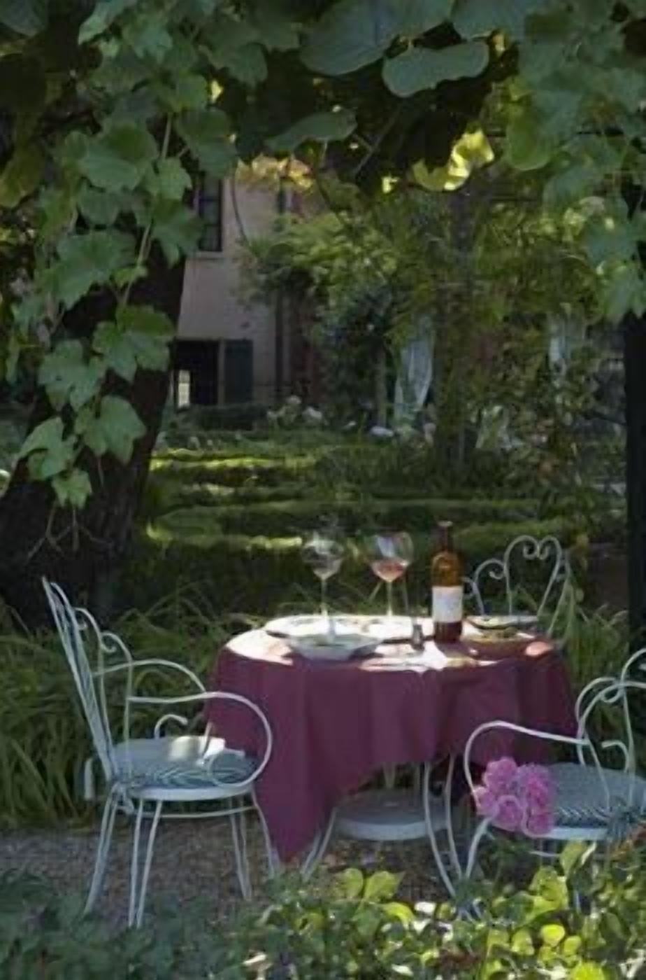 Una mesa redonda con mantel púrpura, rodeada de sillas de metal, ubicada en un jardín con follaje y un edificio al fondo.