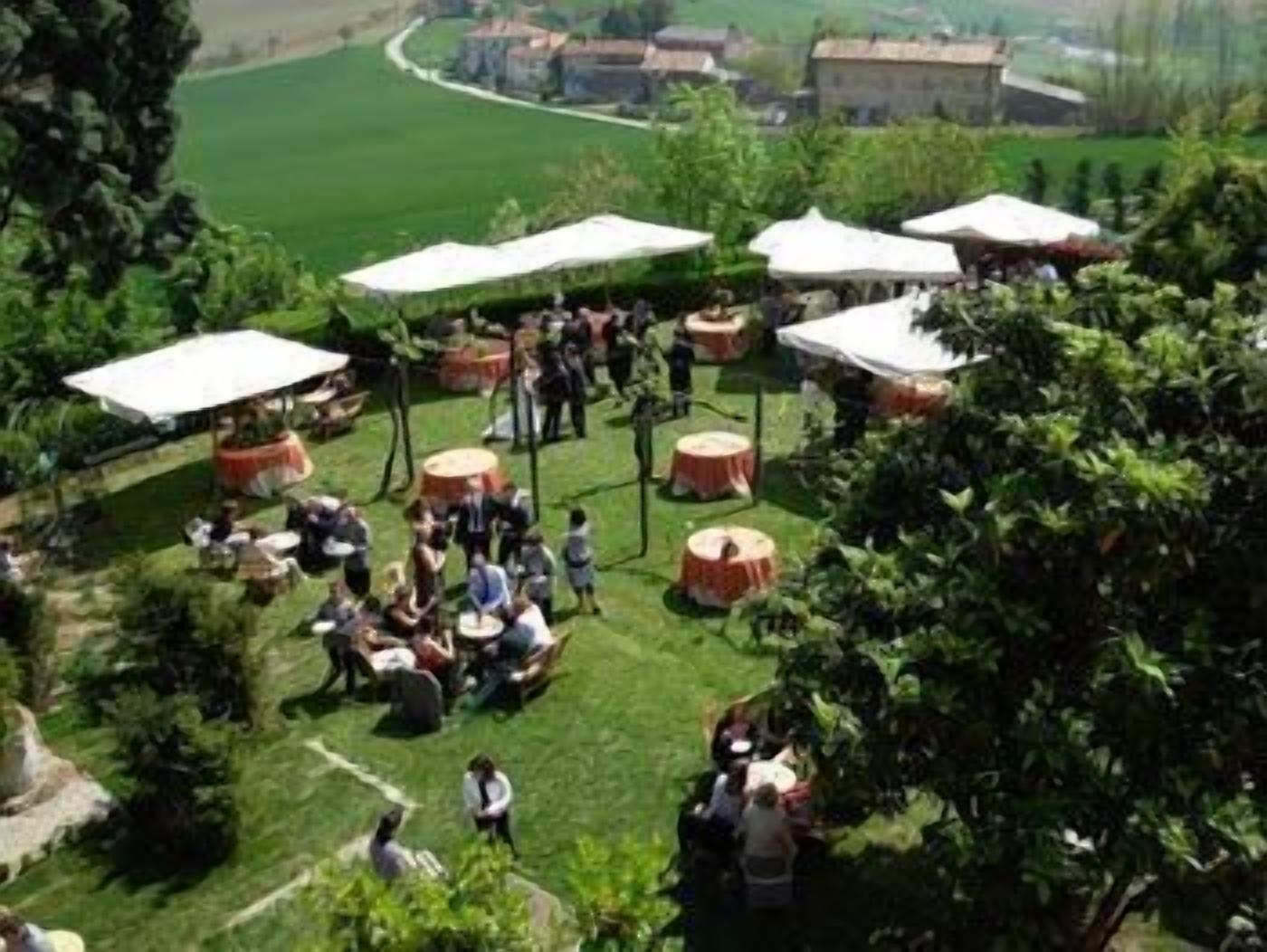 An outdoor event with round tables and white canopies, surrounded by greenery and buildings in the background.