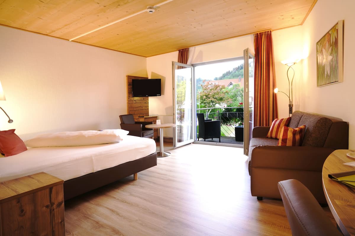A hotel room with a bed, a sofa, a small table, a TV, and a balcony with a view of greenery.