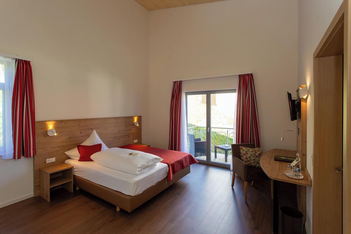 A hotel room with a bed, a small table, a chair, and a balcony with red curtains.