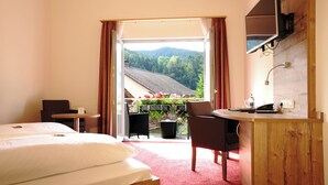 Habitación de hotel con cama, escritorio, silla y televisor. Posee un balcón con vistas a árboles y montañas.
