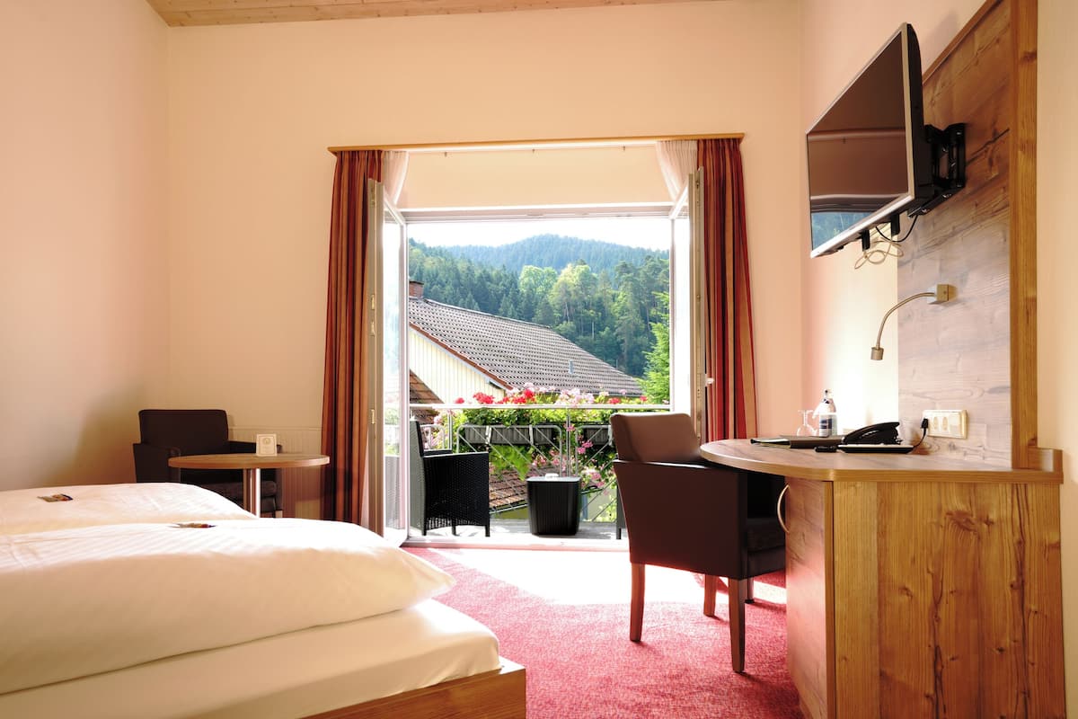 A hotel room with a bed, a desk, a chair, and a TV. There is a balcony with a view of trees and mountains.