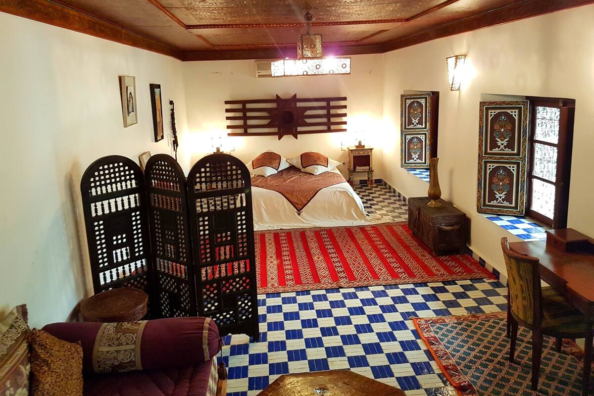 A traditional room with a checkered floor, a bed with a patterned bedspread, a wooden ceiling, and decorative wall panels.
