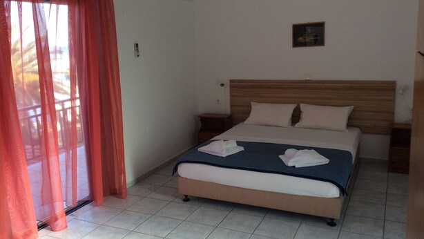 A hotel room with a bed, a nightstand, a small table, and a window with curtains. - Maria's Studios (Sfakia)