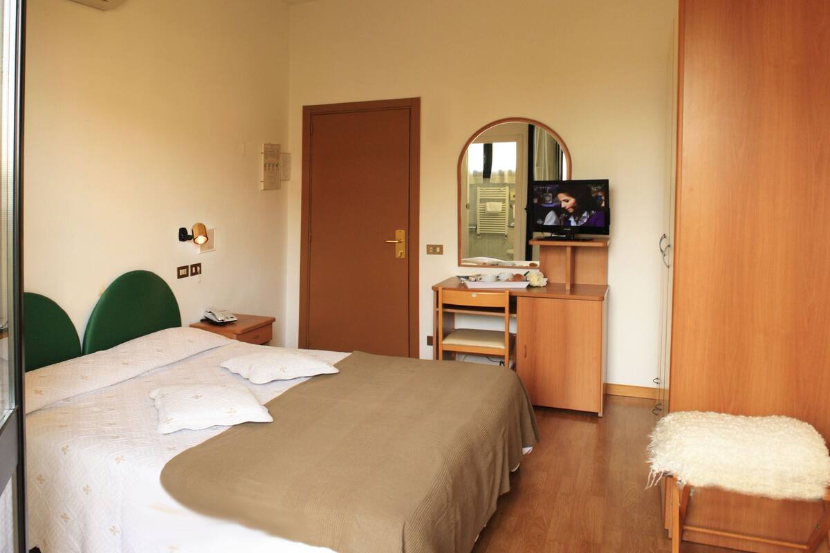 A hotel room with a bed, a desk with a mirror, a TV, and a wooden wardrobe.