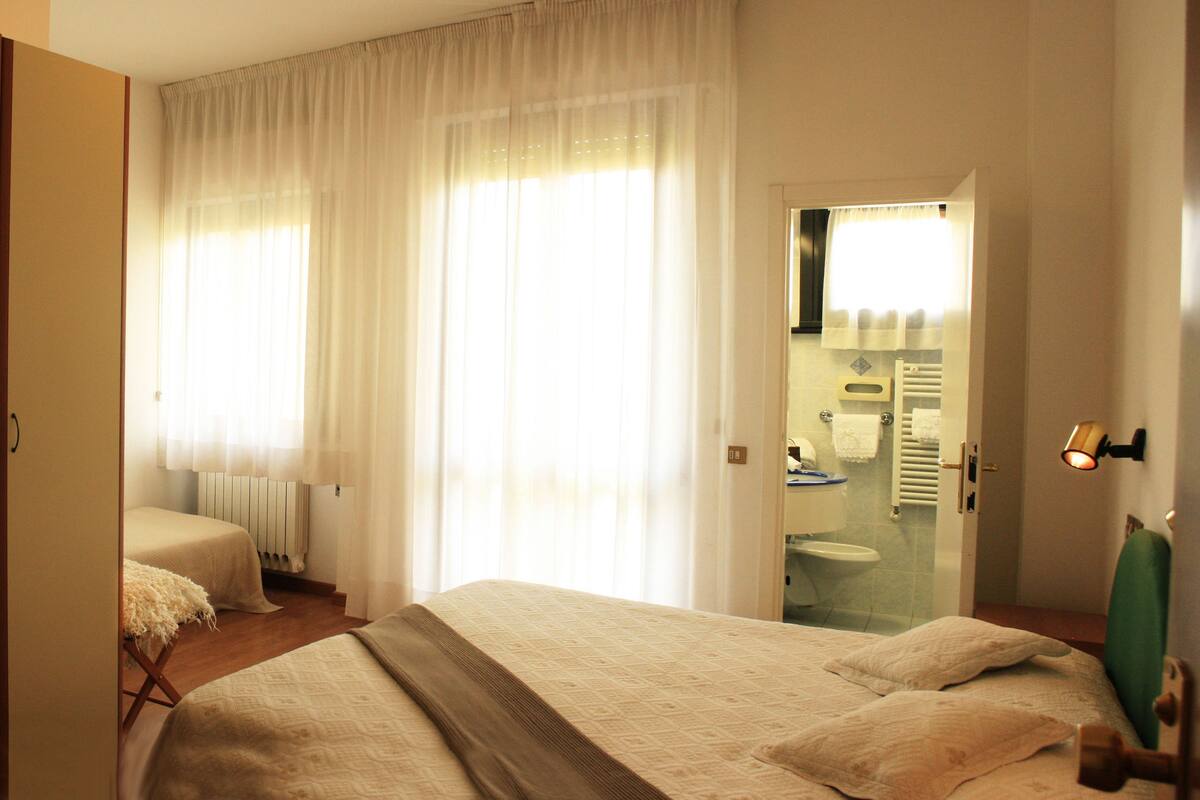 A hotel room with a bed, a mirror, a bathroom visible through a glass door, and a wooden wardrobe.