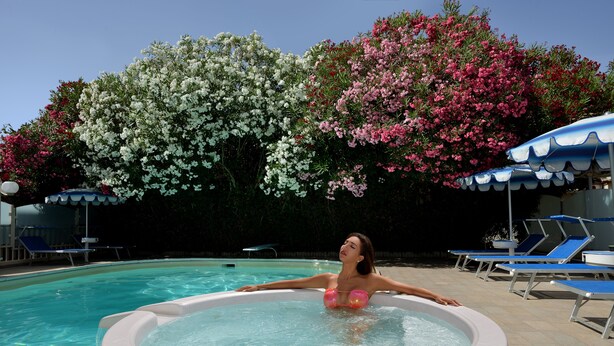 Une femme se détend dans un bain à remous au bord d’une piscine, entourée de chaises longues et de parasols, et bordée d’arbres en fleurs.