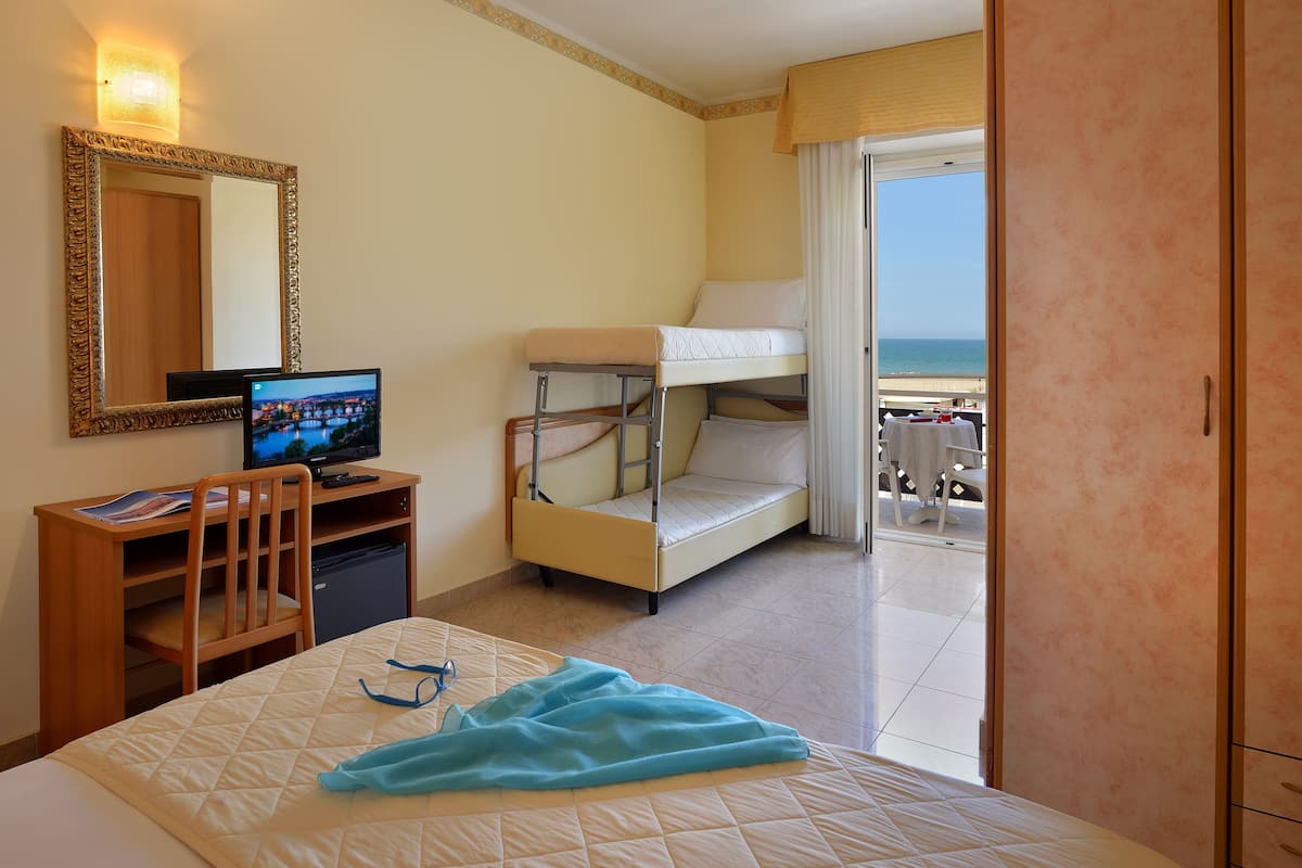 Une chambre d’hôtel avec un lit, un bureau avec un ordinateur, un lit superposé et un balcon donnant sur la mer.
