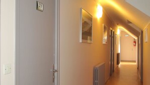 A hallway with a radiator, a door with a number plate, and a light fixture. - Hôtel des Rochers (Marvejols)