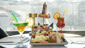 A table with a fruit platter, a martini glass, and a cocktail with a straw, set against a window with a view of buildings. - New Park Hotel Ankara (Ankara)