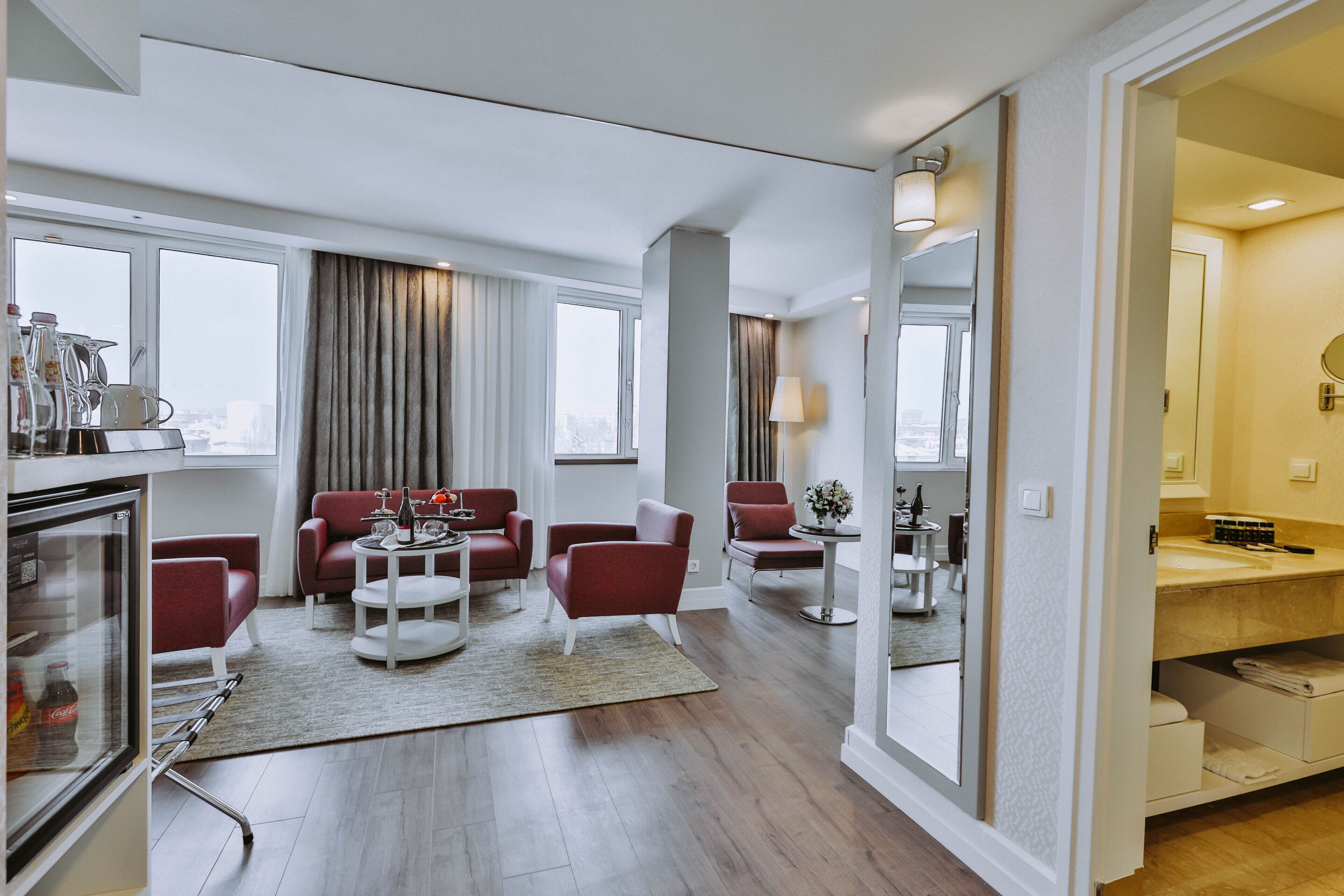 A modern hotel room with a living area featuring red chairs, a round table, and a refrigerator.