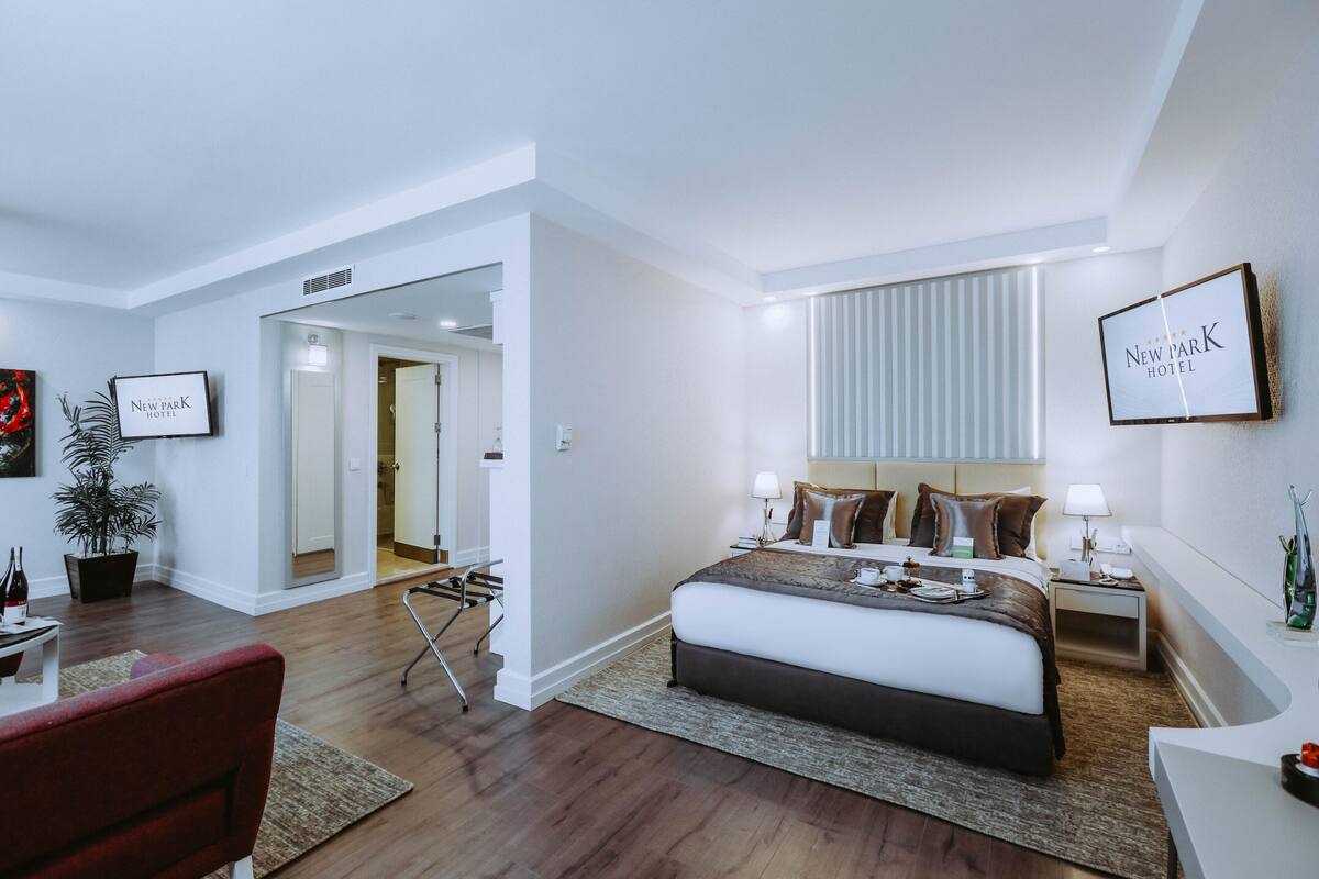 Ein modernes Hotelzimmer mit einem großen Bett, Nachttischen, einem Schreibtisch und einem Sessel. Eine Topfpflanze und ein gerahmtes Schild sind an der Wand zu sehen.
