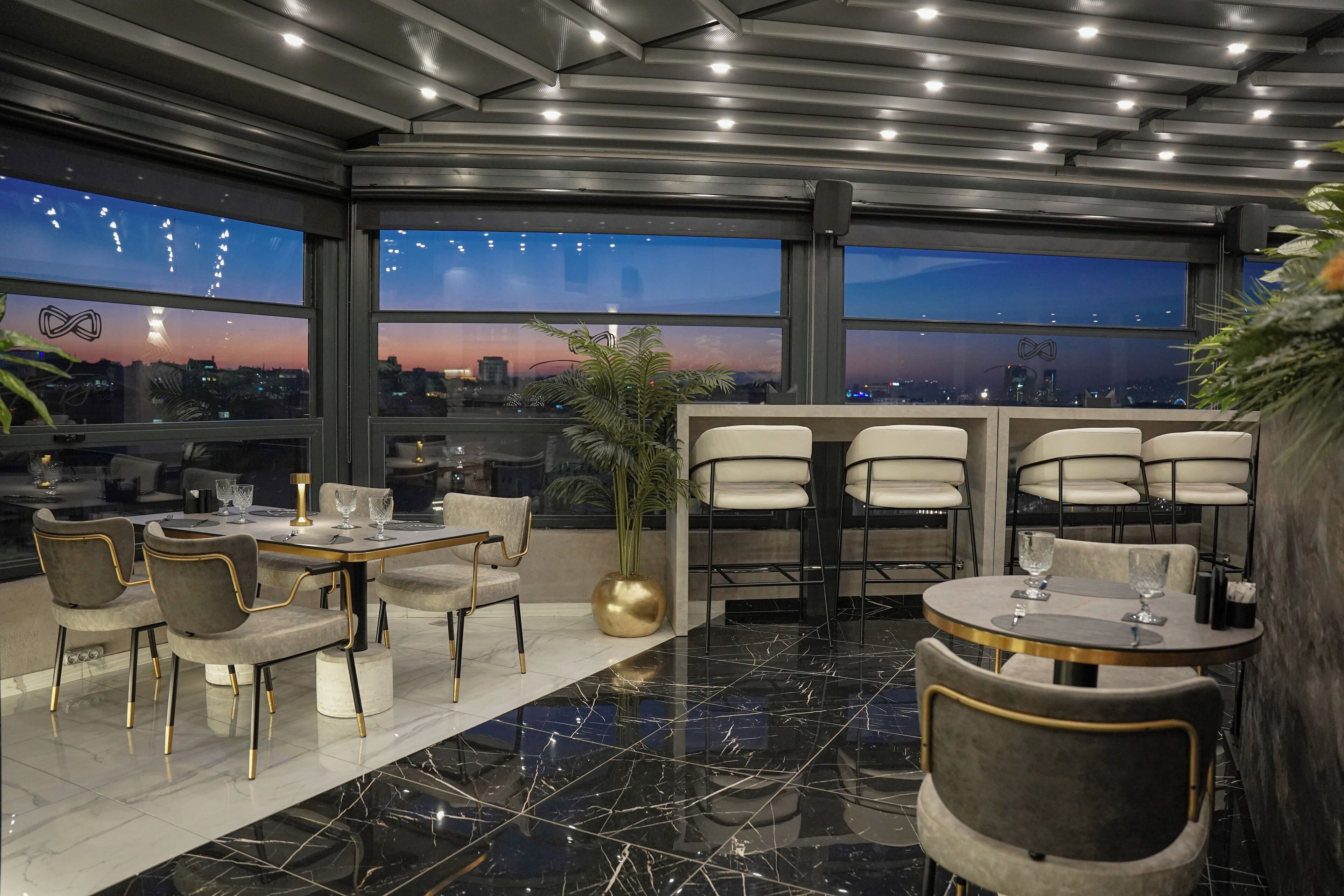 Un comedor moderno con techo alto, ventanas amplias que ofrecen vistas a la ciudad y una mezcla de mesas redondas y rectangulares con sillas.
