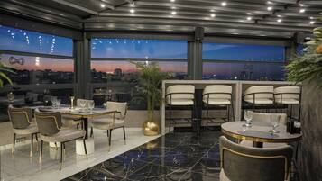 Un comedor moderno con techo alto, ventanas amplias que ofrecen vistas a la ciudad y una mezcla de mesas redondas y rectangulares con sillas.