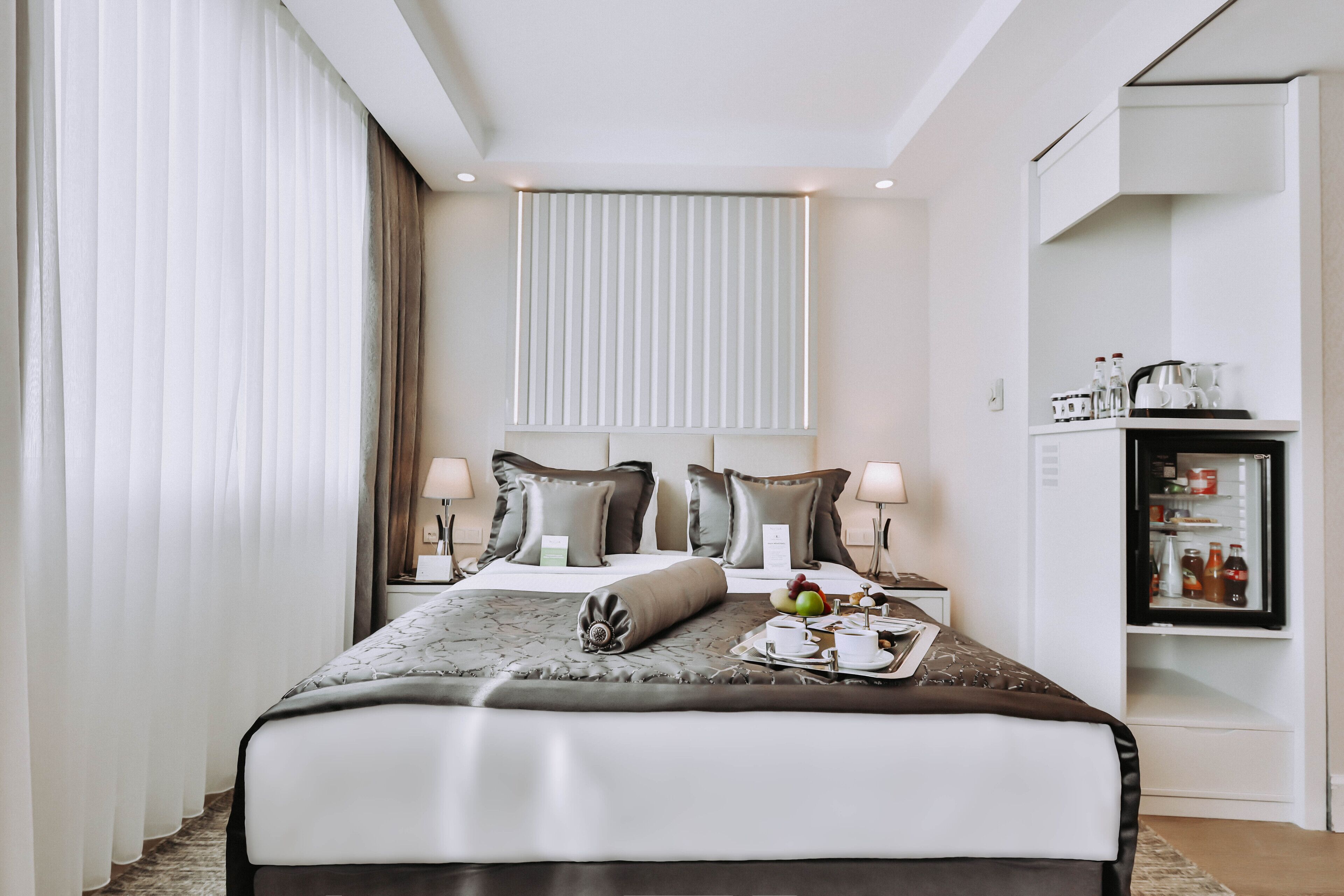 A modern hotel room with a large bed, two bedside tables, a minibar, and a view of the city.