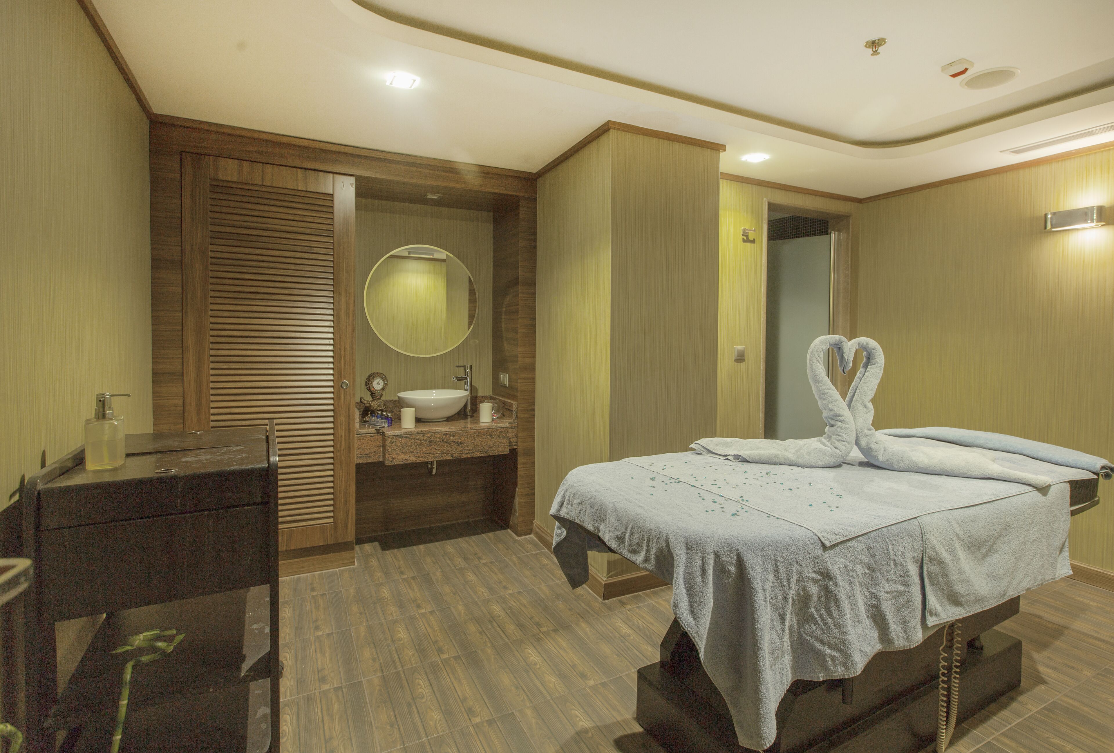A hotel room with a wooden vanity, a round mirror, a bed with towels, and a small table with a bottle and a cup.