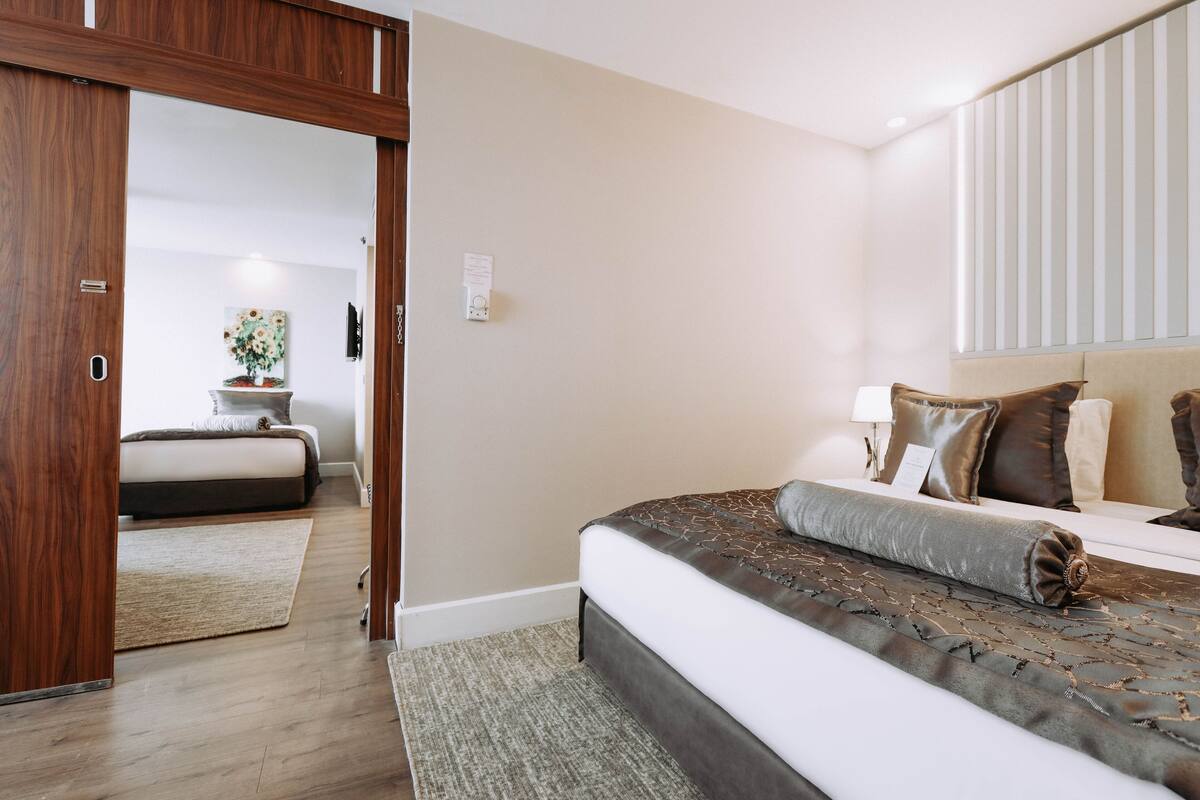 Ein Schlafzimmer mit einem großen Bett, einer Holztür, einem kleinen Tisch mit einer Vase und einem Fenster mit Jalousien.