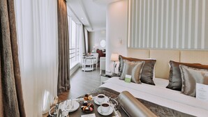 Habitación de hotel con cama, una mesa de comedor con una bandeja de pasteles y café, un escritorio con lámpara y vistas a la ciudad.