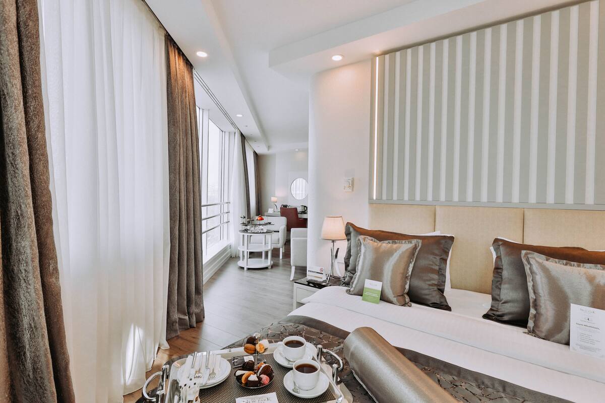 Ein Hotelzimmer mit Bett, Esstisch mit einer Auswahl an Gebäck und Kaffee, Schreibtisch mit Lampe und Blick auf die Stadt.