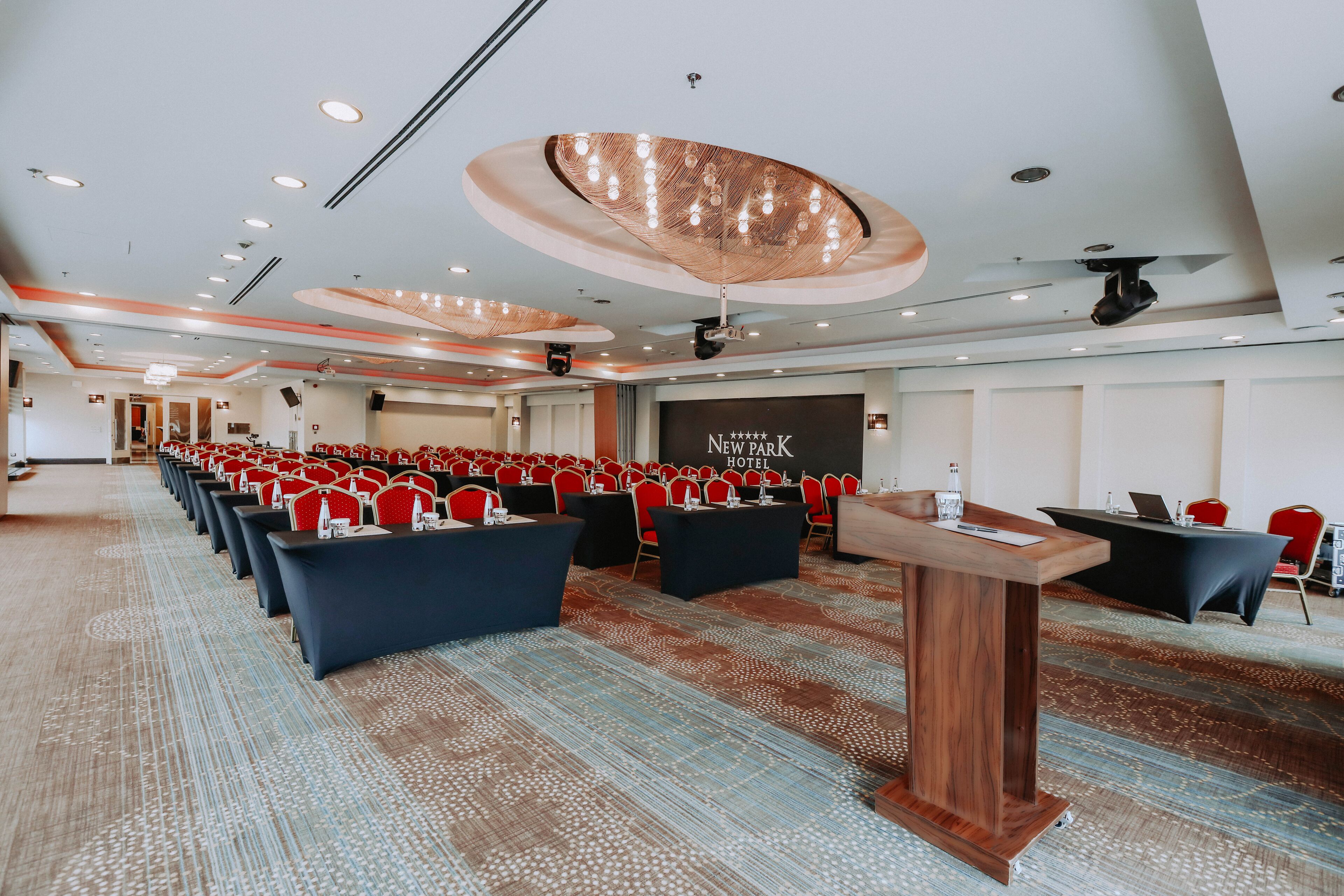 Ein Konferenzraum mit Reihen roter Stühle, einer Bühne mit Rednerpult und einem Banner mit der Aufschrift 'New Park Hotel'.