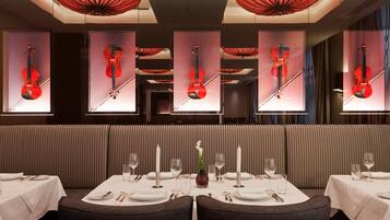 A restaurant with a modern interior, featuring a long dining table set for a meal, a vase with a single white flower, and red violin-themed wall decorations.