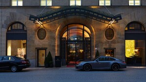 A historic building with a grand entrance, two cars parked in front, and a cobblestone street.