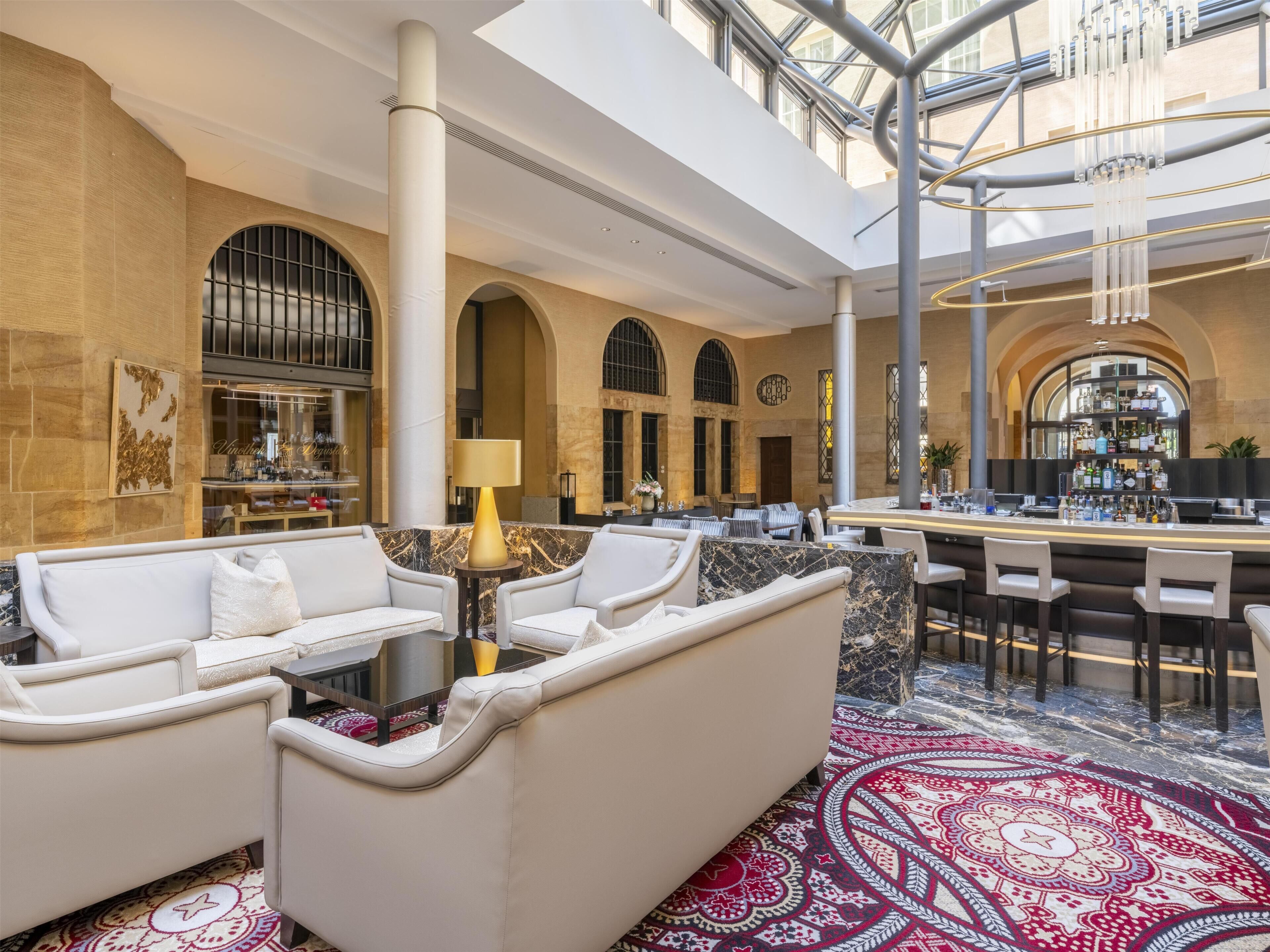 A spacious lobby with a high ceiling, marble flooring, and a central seating area with white sofas and a patterned rug.