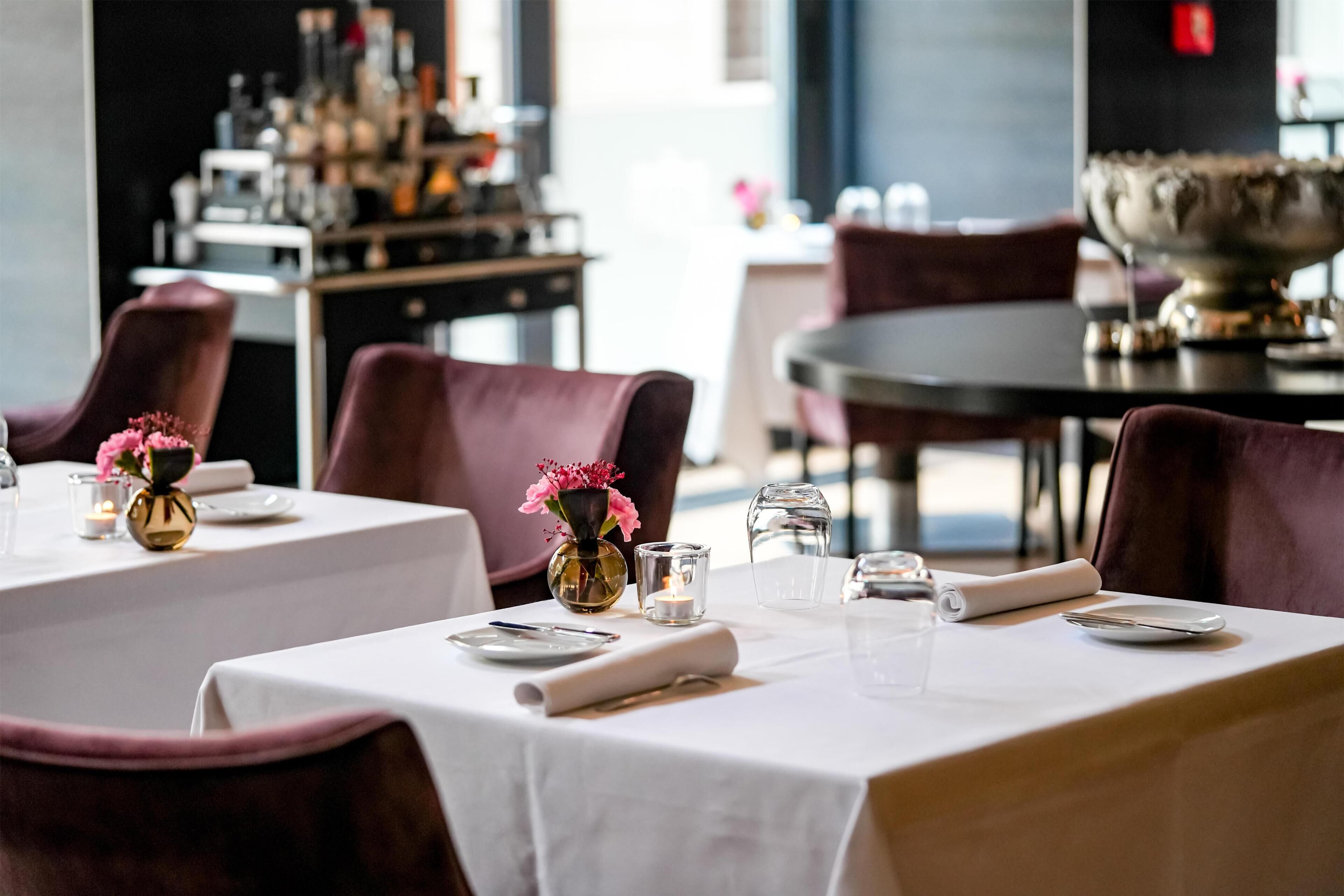 A restaurant with tables set for dining, featuring white tablecloths, vases with flowers, and neatly arranged cutlery.