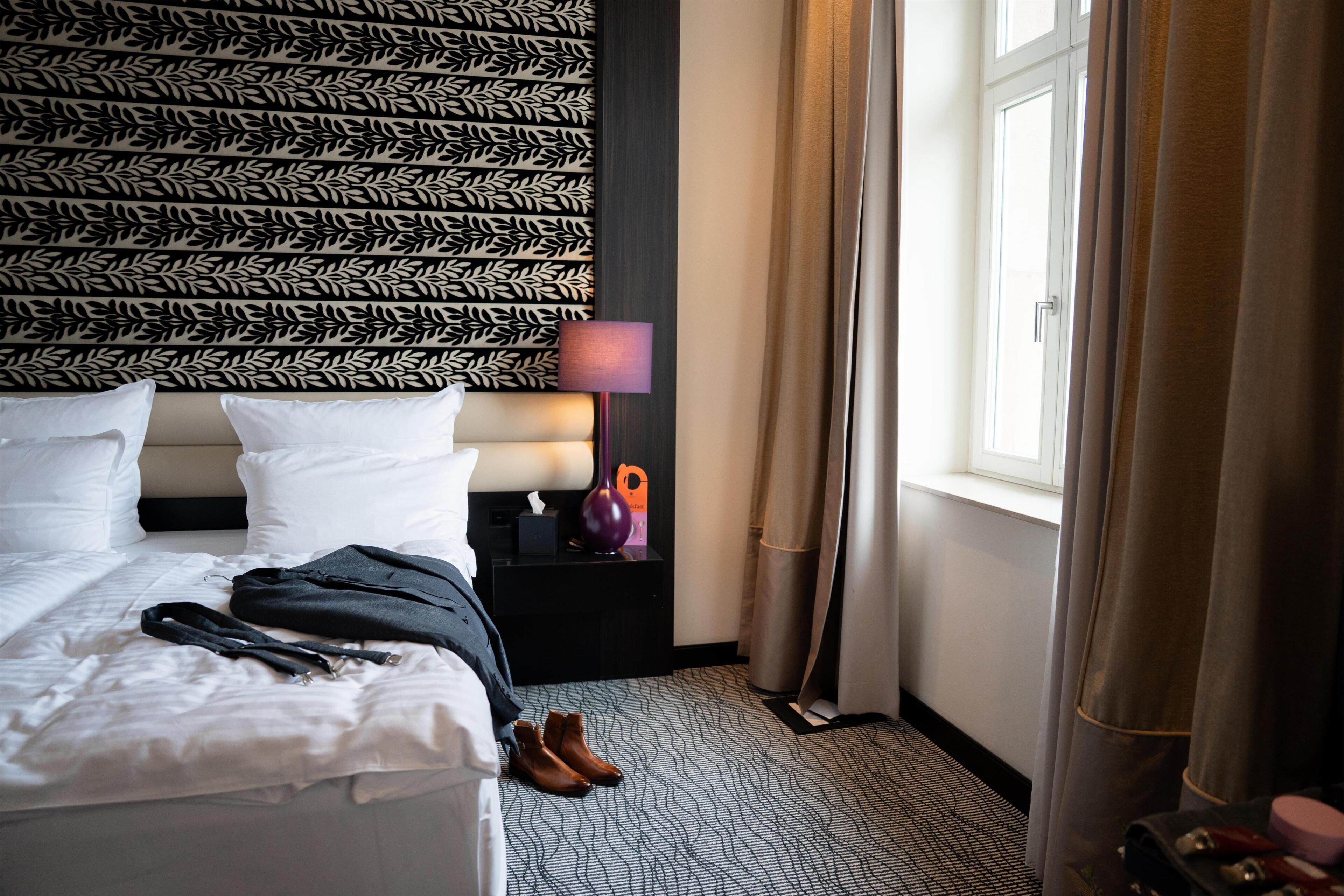 A hotel room with a patterned headboard, a bed with white linens, a bedside table with a lamp, and a window with curtains.