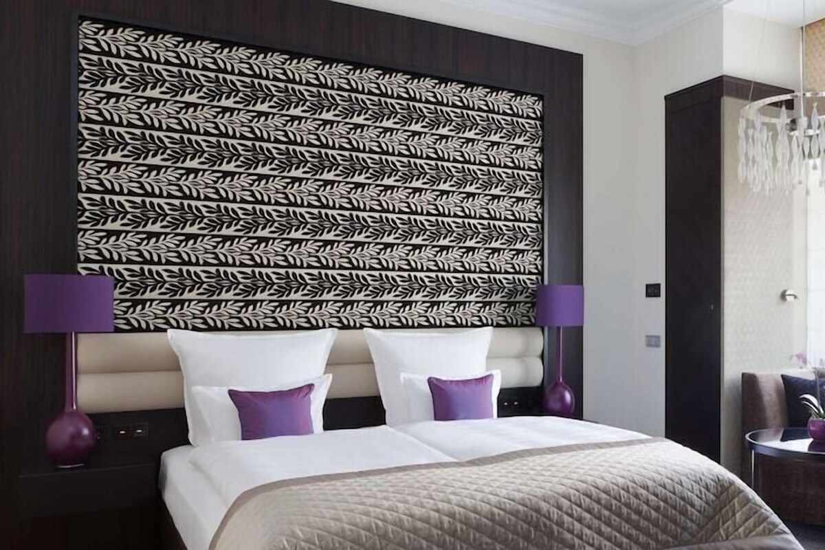A modern hotel room with a large bed, two nightstands, a chandelier, a sofa, and a window with a view.