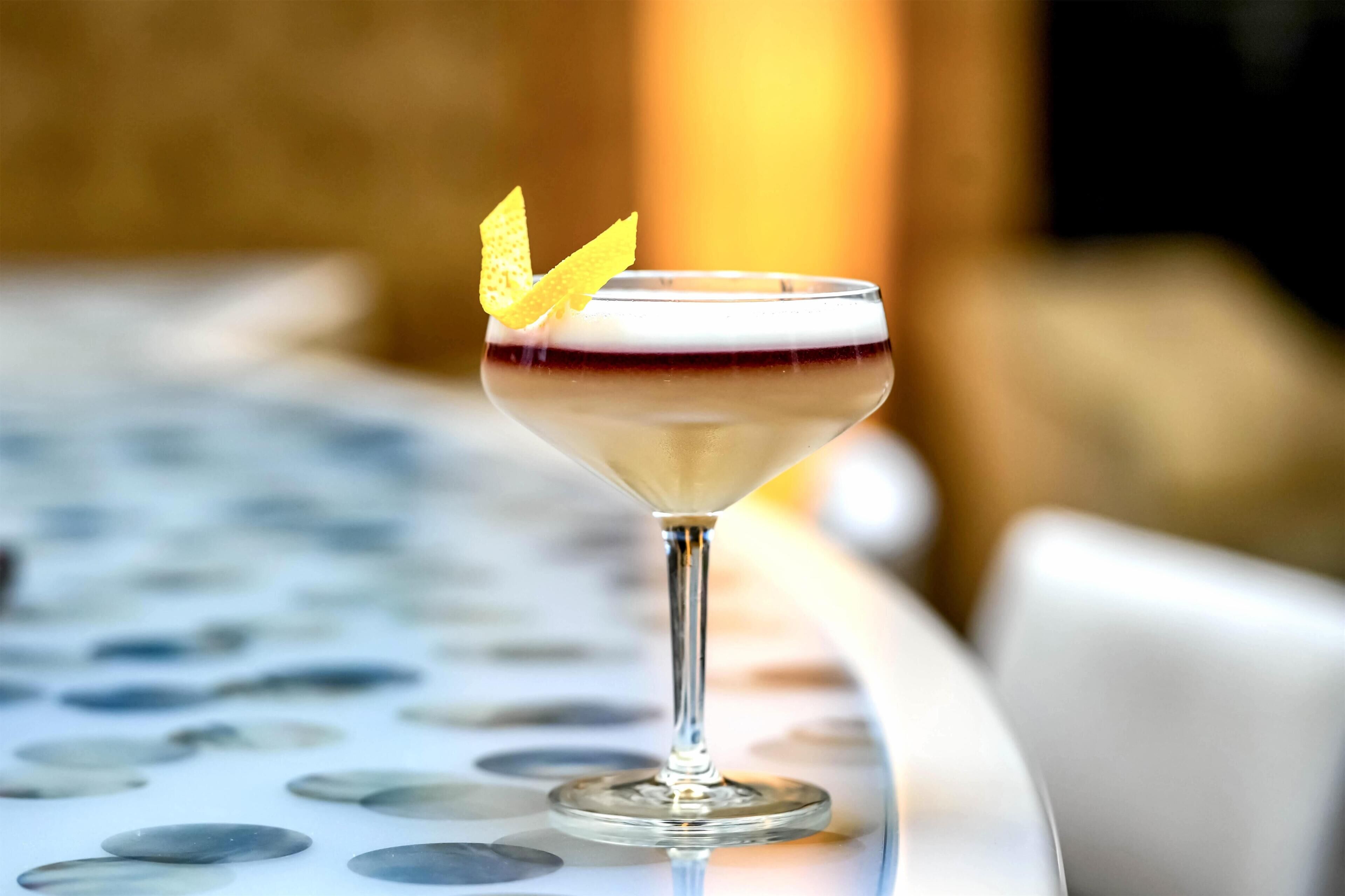 Un cóctel en una copa de martini, adornado con una rodaja de limón, sobre una mesa con superficie reflectante.