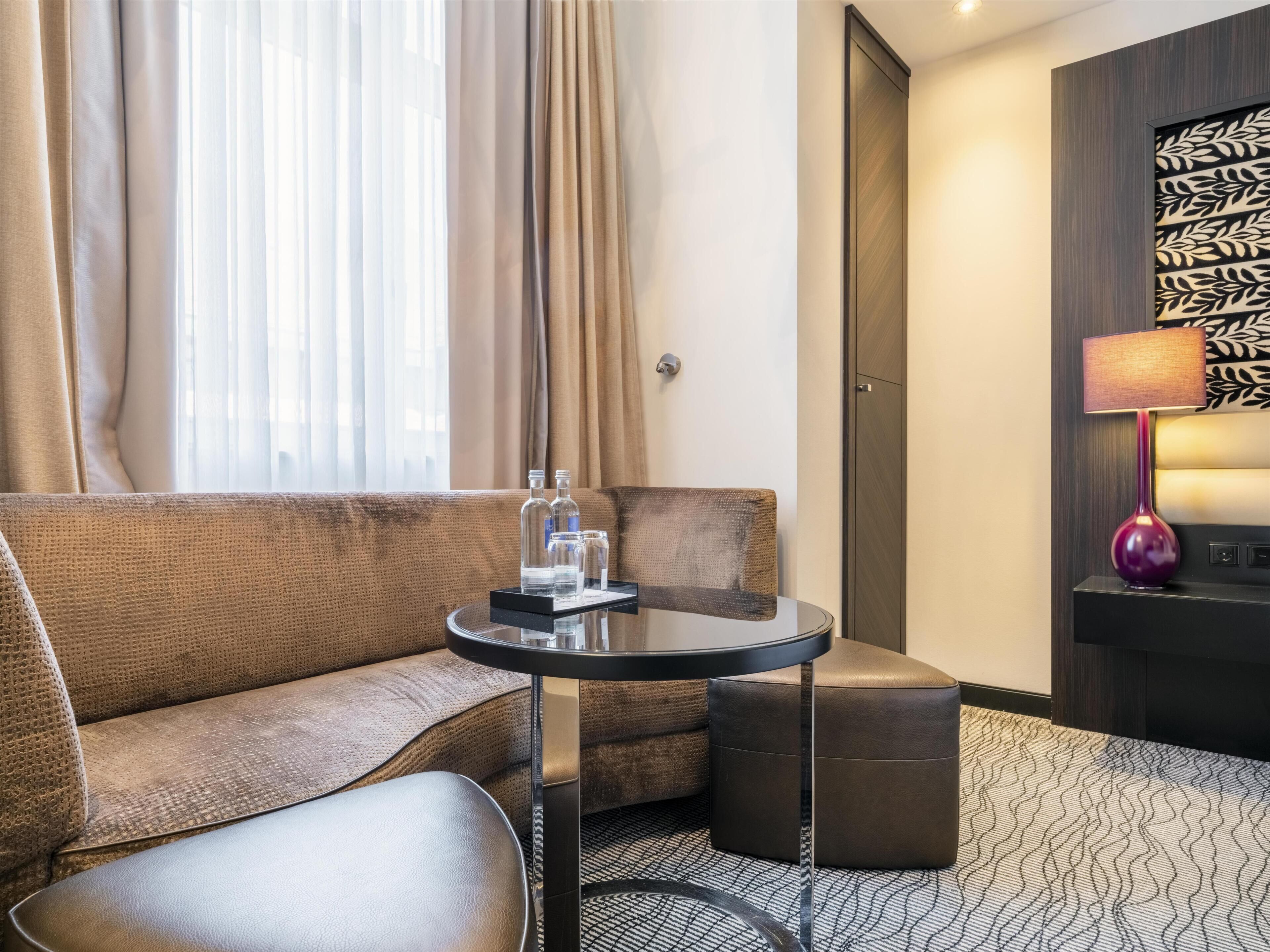 A hotel room with a sofa, a small round table, a lamp, and a patterned carpet.