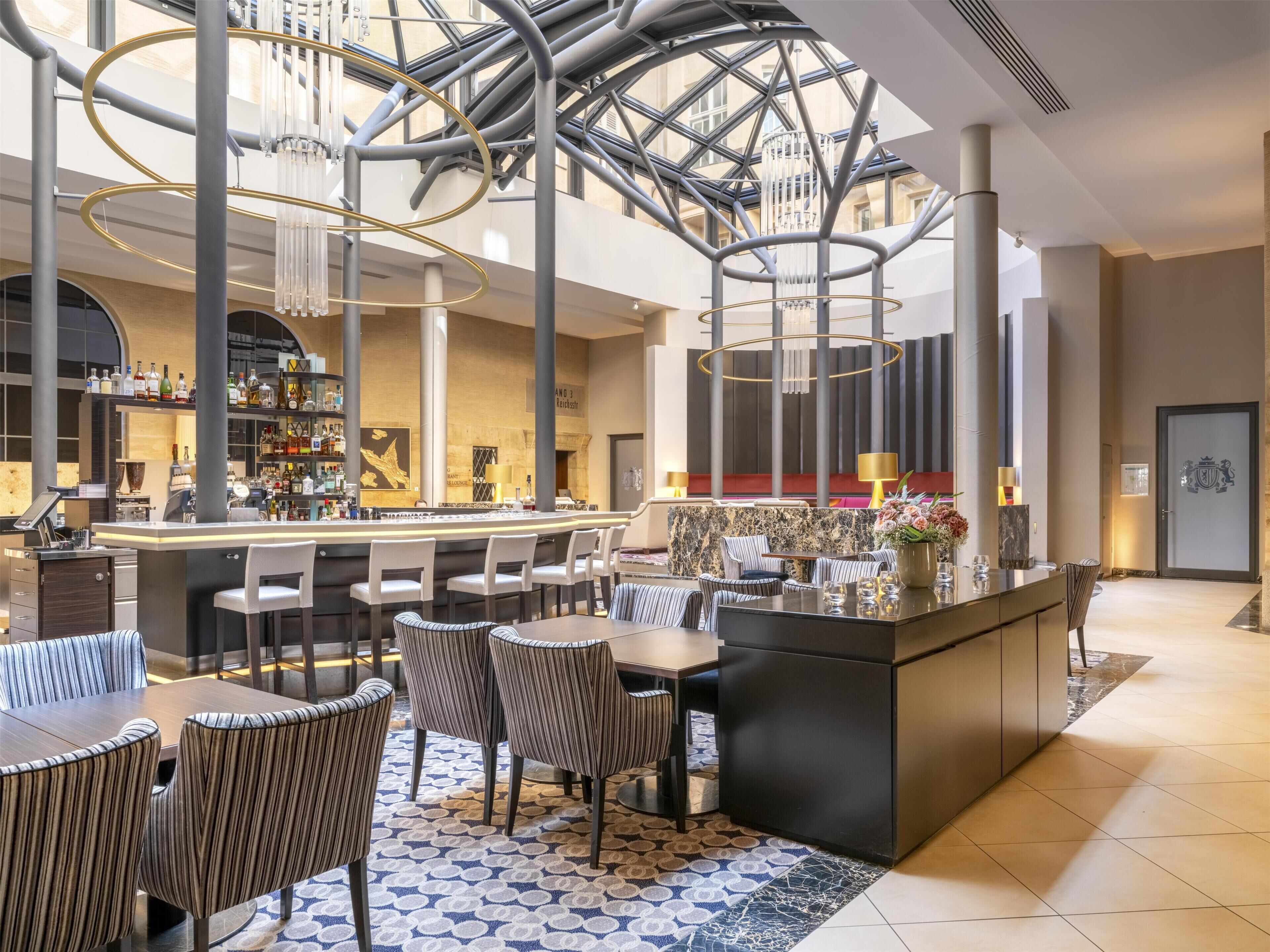 A modern hotel lobby with a bar area, seating arrangements, and a high ceiling with exposed structural elements.