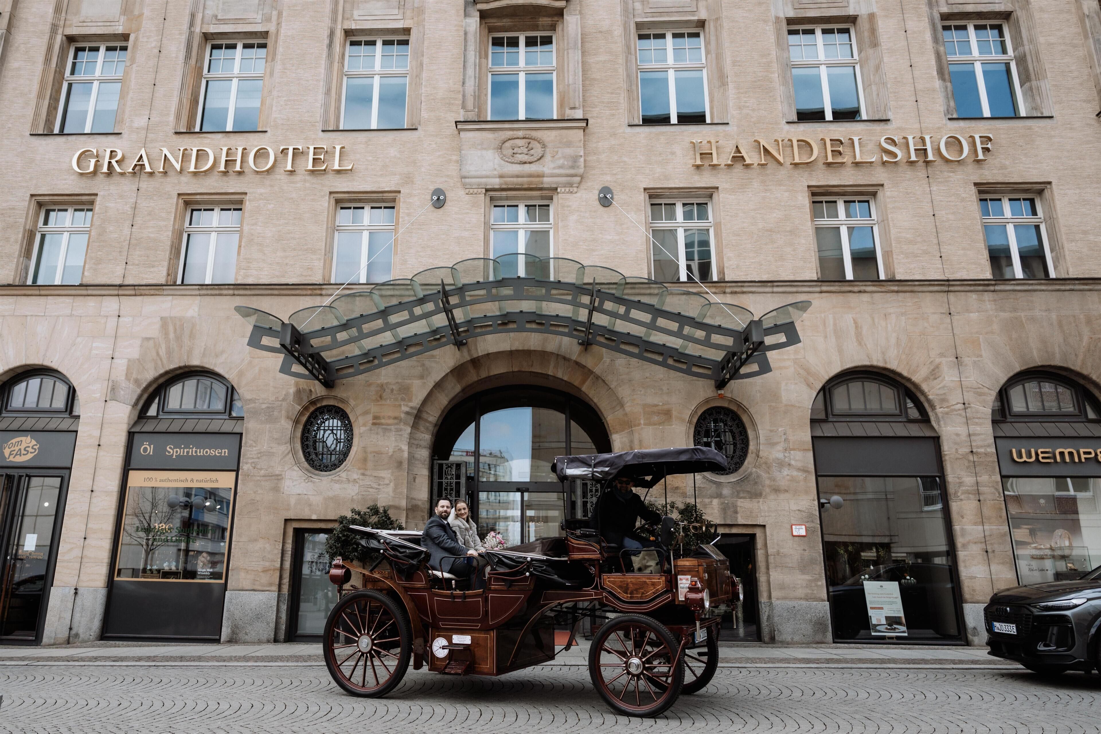 Ein prächtiges Gebäude mit den Schildern 'GRANDHOTEL' und 'HANDELSHOP', eine Kutsche und ein Auto vor der Fassade.