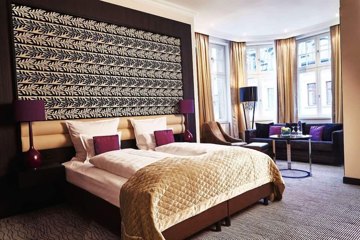 Une chambre d’hôtel avec un grand lit, une tête de lit ornée d’un motif de feuilles, un bureau avec une lampe, une chaise et un canapé.