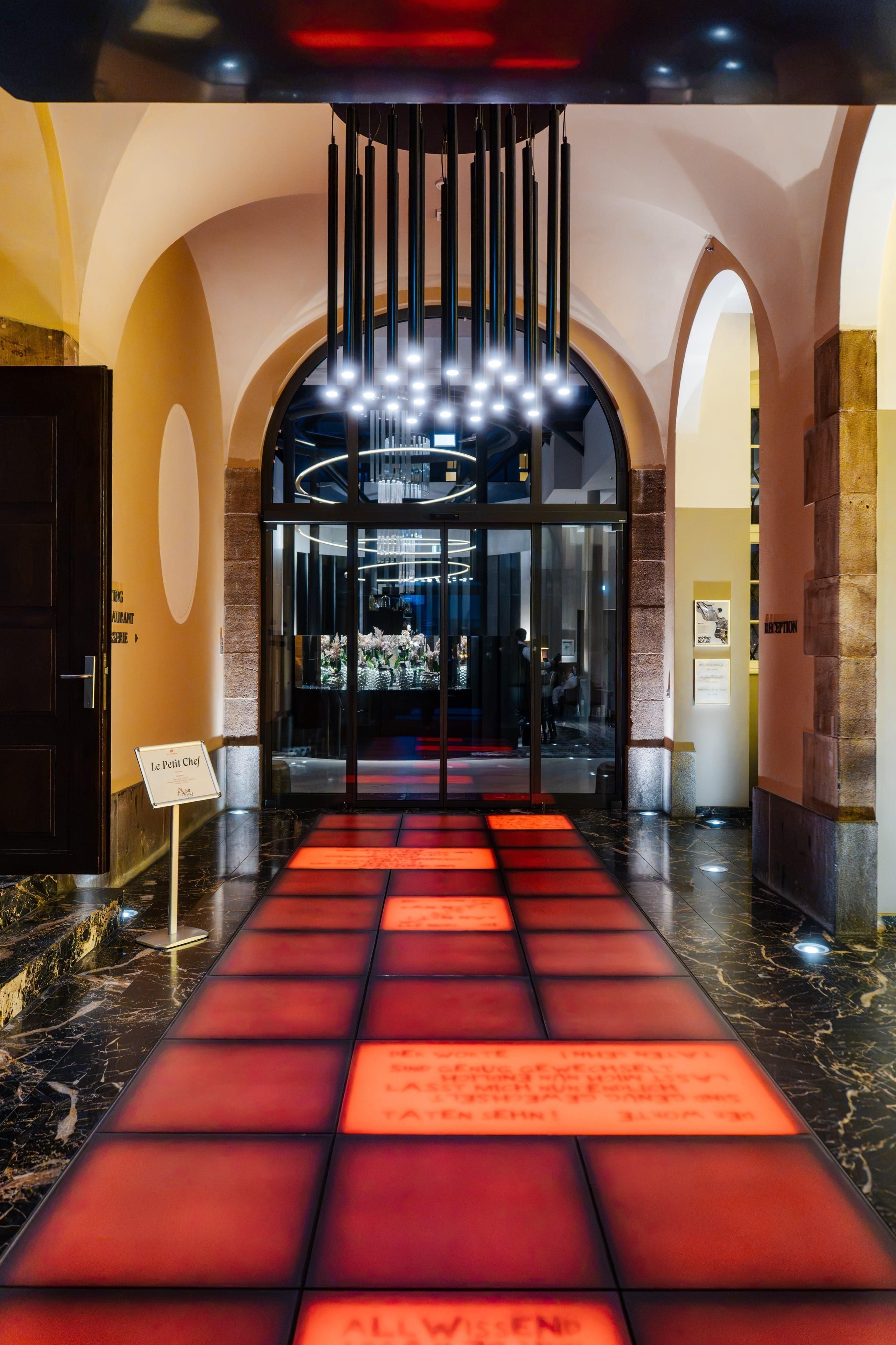 Un hall de hotel de diseño moderno, con entrada de cristal, un camino iluminado con luz roja y un letrero que indica el restaurante.