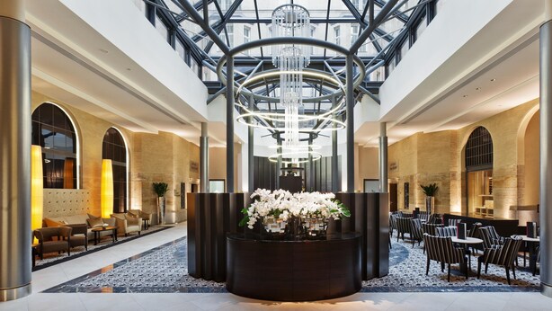 A spacious lobby with a central reception desk, seating areas, and a high ceiling with a modern chandelier.