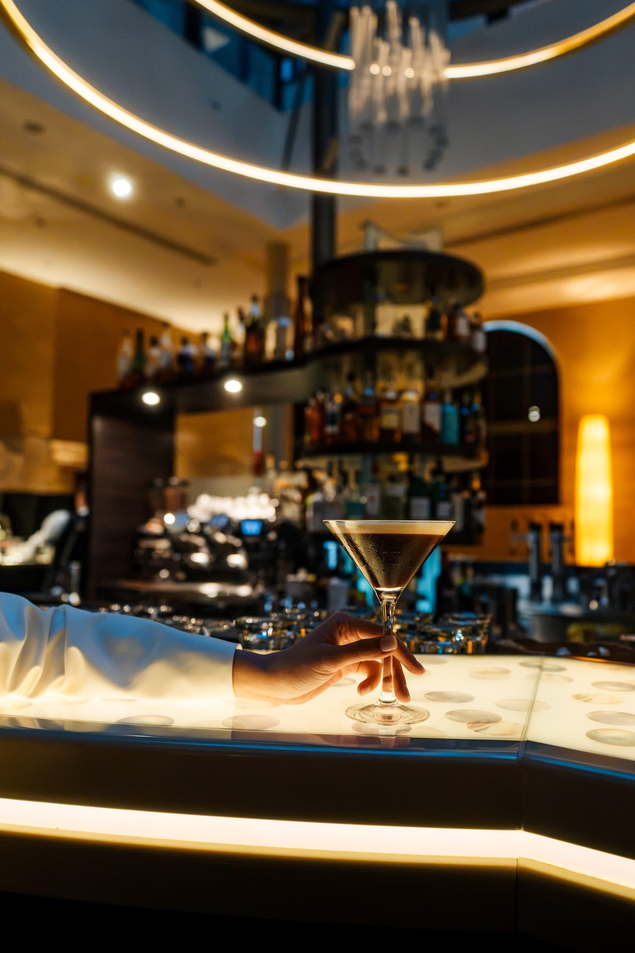 Zona de bar con una mano sosteniendo una copa de martini, una barra y estantes llenos de botellas.