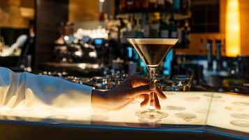 Zona de bar con una mano sosteniendo una copa de martini, una barra y estantes llenos de botellas.