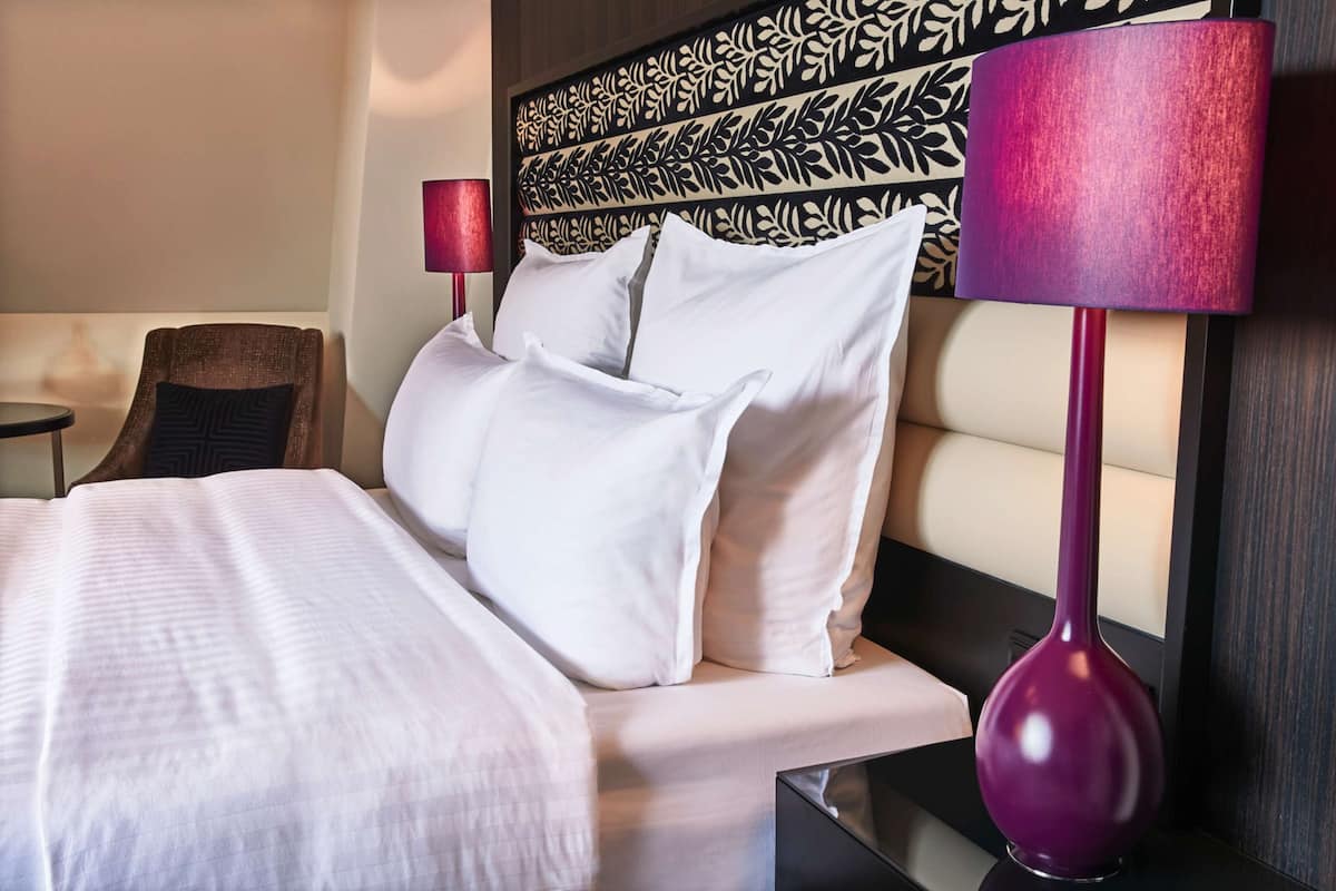 Un lit bien fait, avec du linge de lit blanc, une tête de lit moderne ornée d’un motif de feuilles, une lampe rouge et un vase violet posé sur une table de chevet.