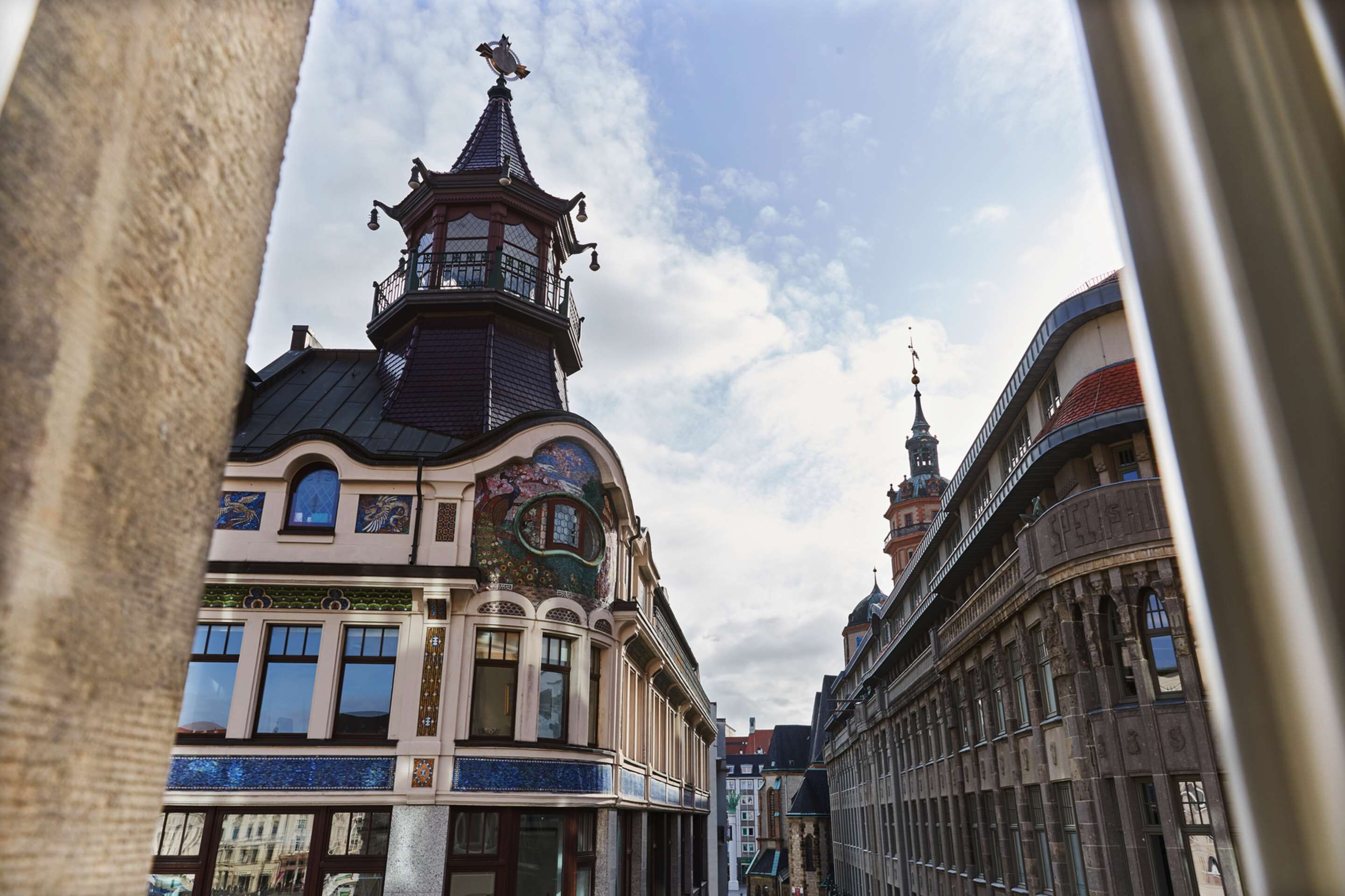 A street view with a building featuring a tower with a spire and a balcony.