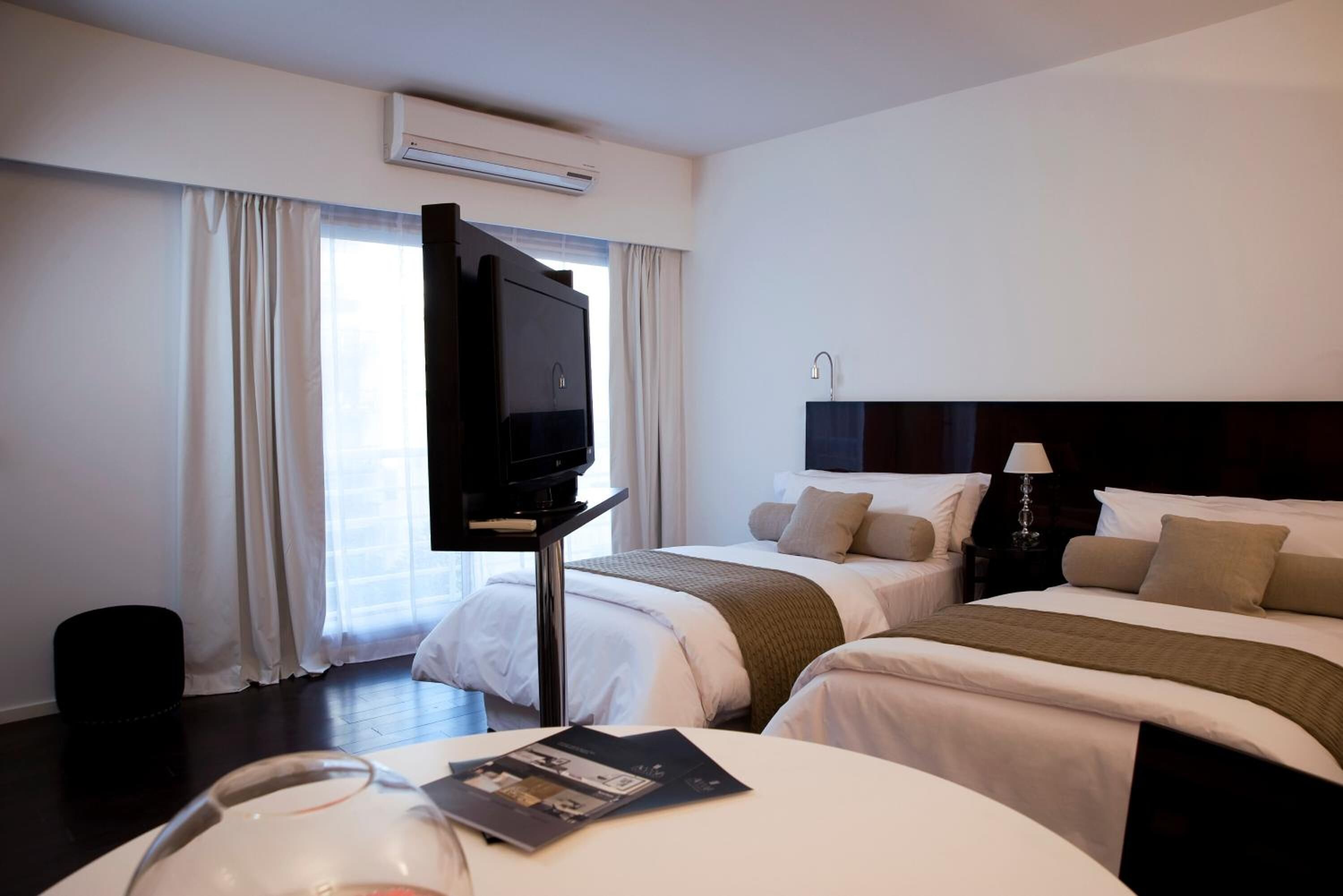 Habitació d'hotel amb dos llits, un televisor sobre un suport, una taula de nit amb una làmpada i una finestra amb cortines.
