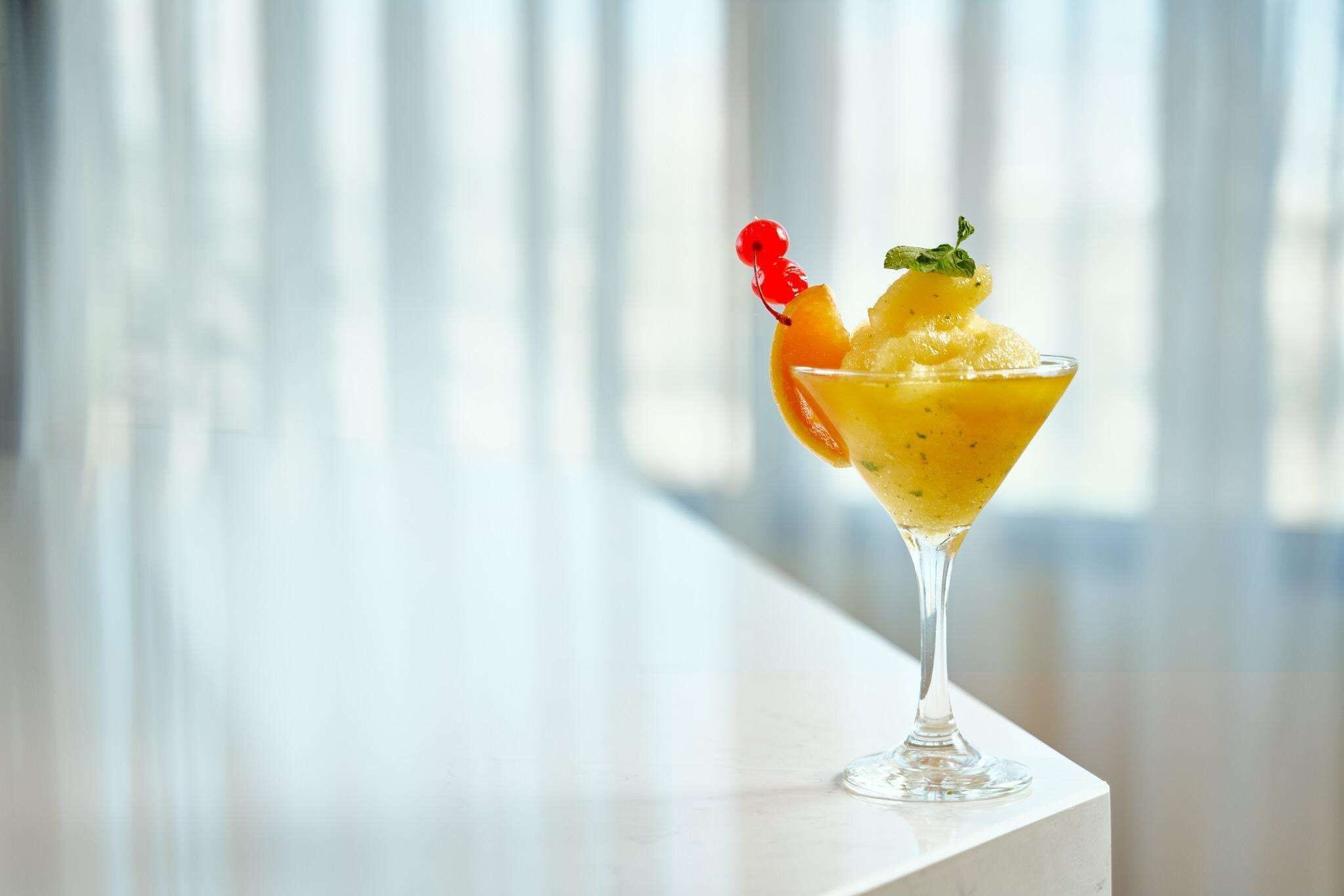 En cocktail i ett martiniglas, garnerad med en dekoration, placerad på en vit yta med ett fönster i bakgrunden.