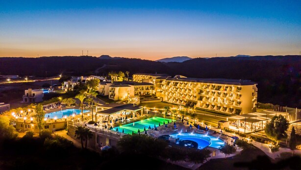 A large hotel with multiple swimming pools, a well-lit outdoor area, and a view of the surrounding landscape.