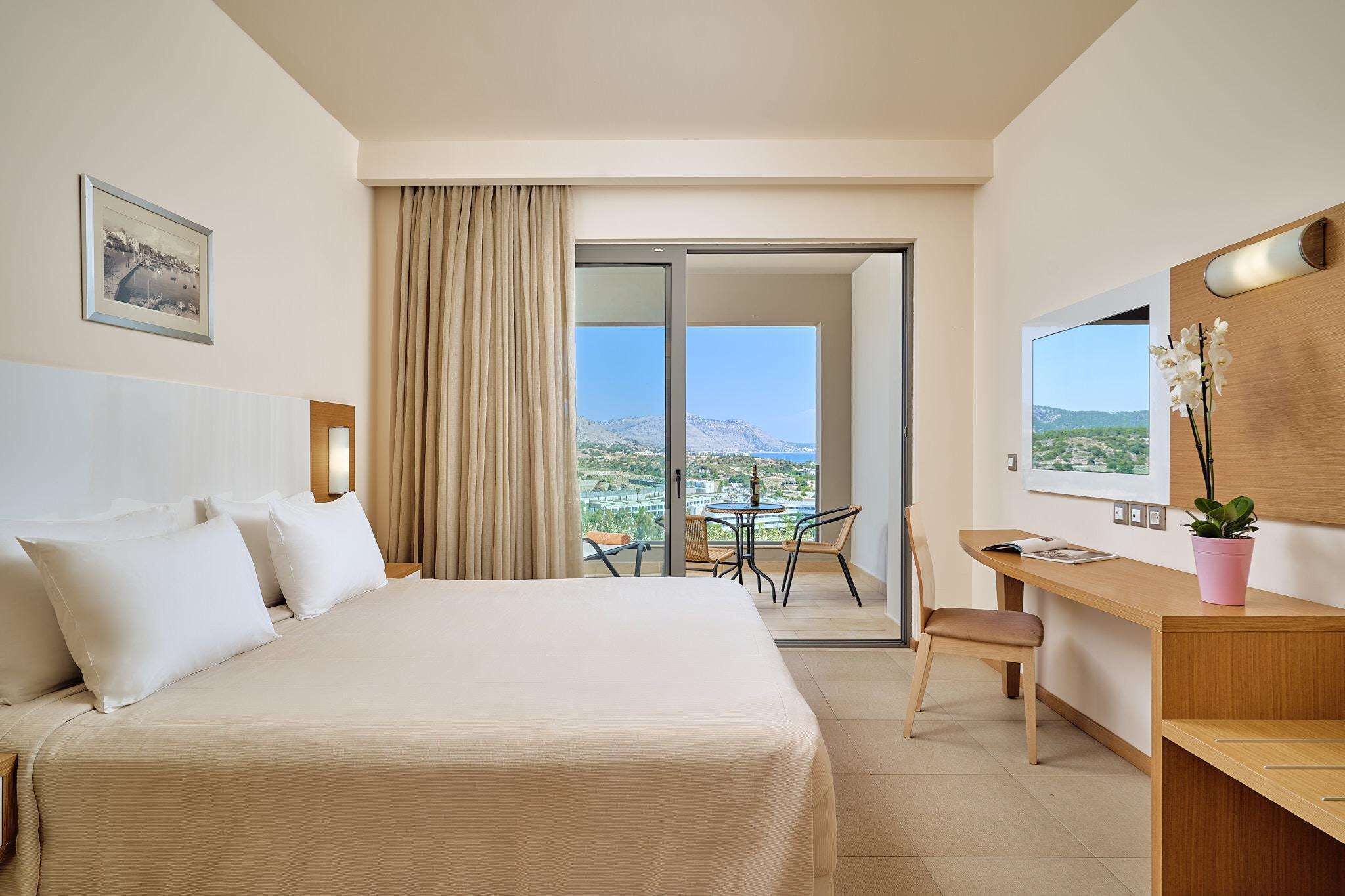 A hotel room with a bed, a desk, a chair, a small table, a potted plant, and a view of the landscape.
