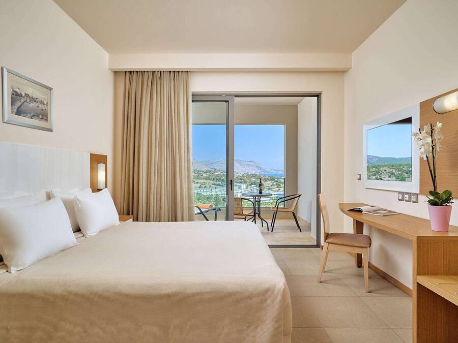 A hotel room with a bed, a desk, a chair, a small table, a potted plant, and a view of the landscape.