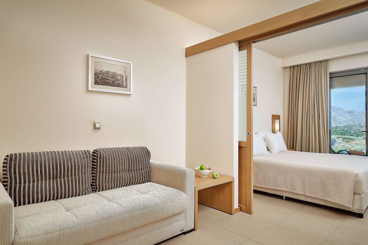 A hotel room with a sofa, a bed, a small table with apples, and a cityscape view through the window.