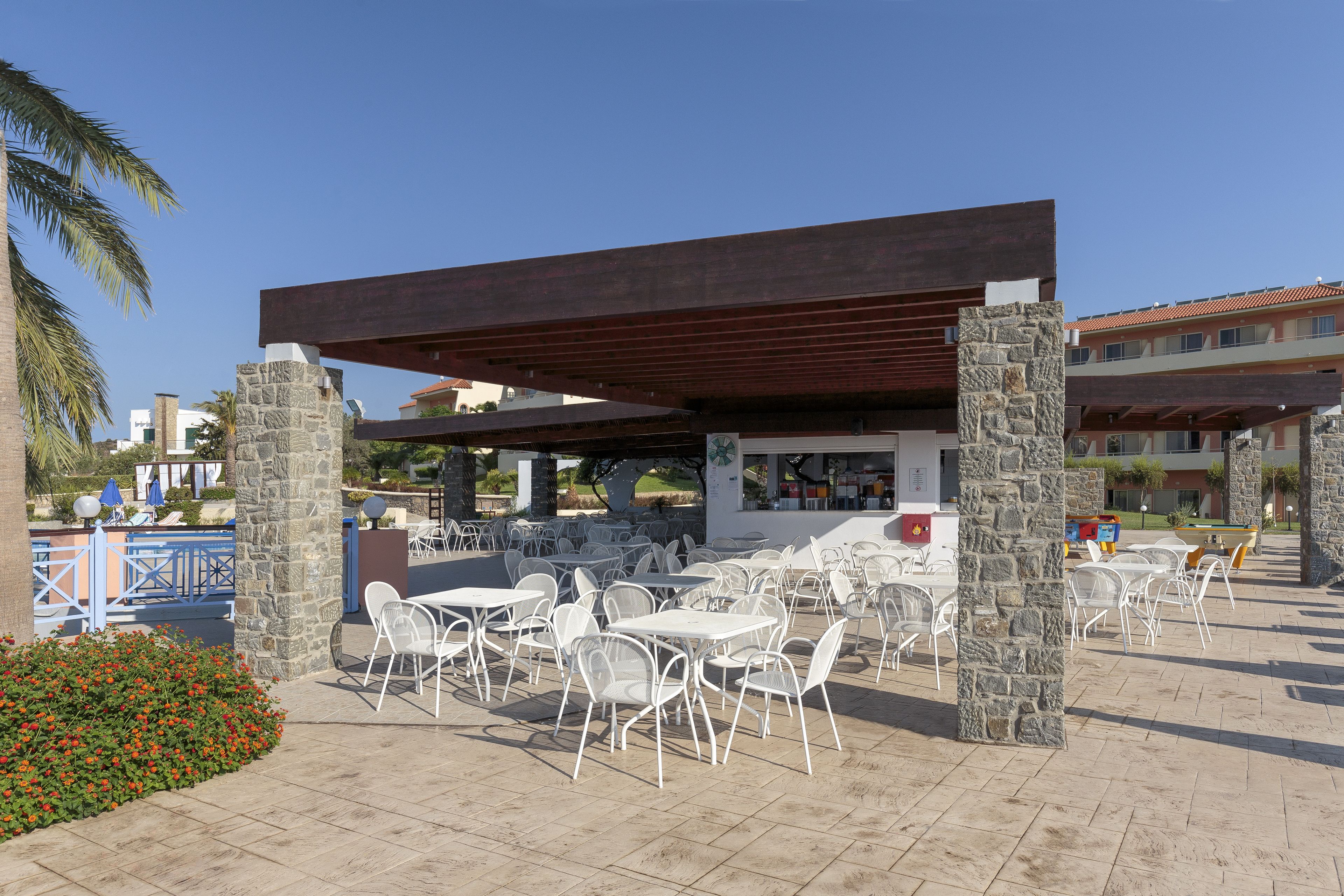 Un área de café al aire libre con pilares de piedra, mesas y sillas de metal blanco, y una zona de estar cubierta.